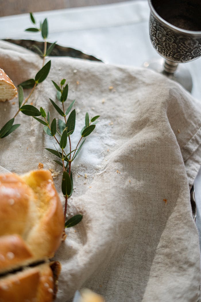 creative-01 A close-up of challah bread, greenery, and a silver cup on a linen cloth.