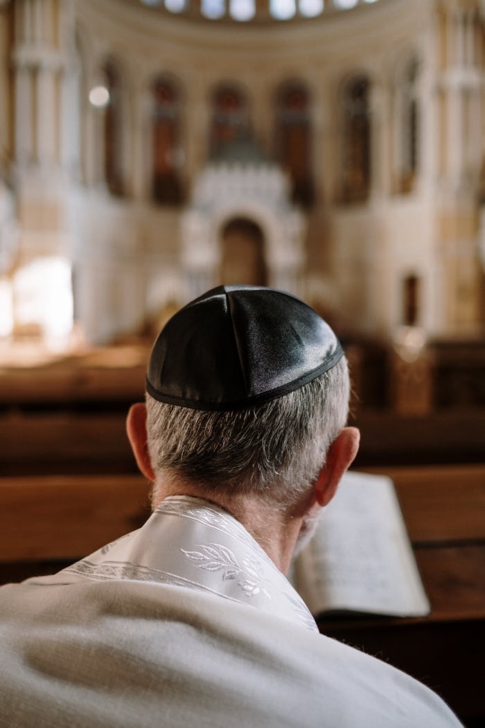 brand-02 A man with kippah is sitting in a synagogue, viewed from the back, reading.
